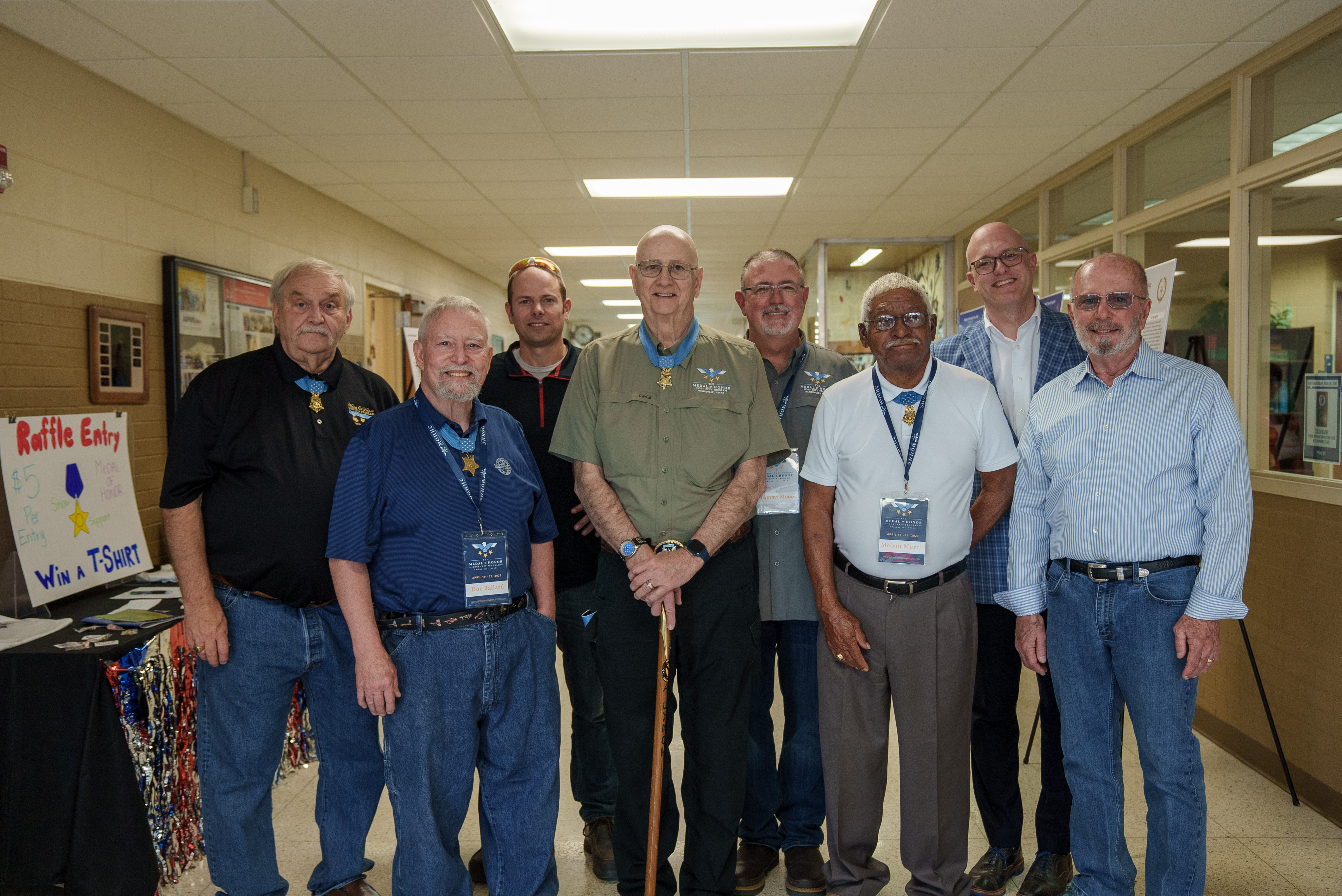 A group photo includes 7 medal of honor recipients and GAF employee Drew Daniel. They are all smiling in a hallway at a medal of honor event in Gainesville, Texas.