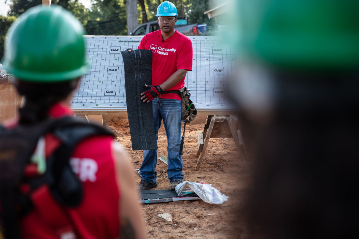 A GAF instructor in a read shirt holds roofing material in front of a group of GAF volunteers at a Habitat build site.